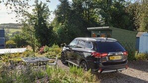 Un SUV noir est garé dans un jardin où se trouvent une table et des chaises, le tout étant entouré de verdure et d’un petit abri de jardin.