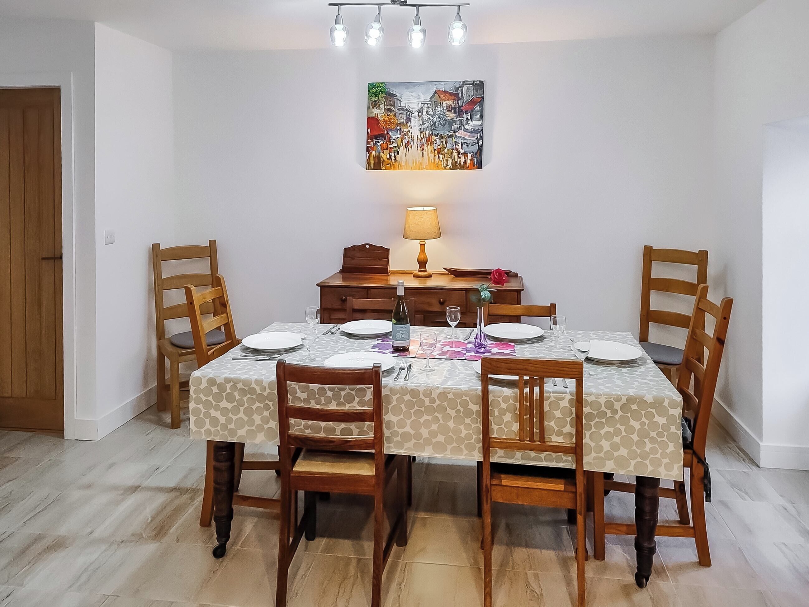 Un coin repas avec une table dressée pour quatre personnes, des chaises en bois, une lampe et un tableau accroché au mur.
