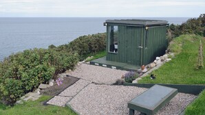 A small, green wooden structure with a door and windows, situated on a gravel path with a grassy area and shrubs on one side and a body of water on the other.