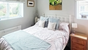 Un lit bien fait, avec une tête de lit blanche et plusieurs oreillers, une table de chevet avec une lampe, et une fenêtre donnant sur un paysage verdoyant.