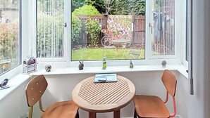 Une pièce dotée d’une grande fenêtre en saillie donnant sur un jardin. On y trouve une table ronde avec deux chaises, une petite étagère ornée d’objets décoratifs et un rebord de fenêtre sur lequel sont posées quelques plantes.