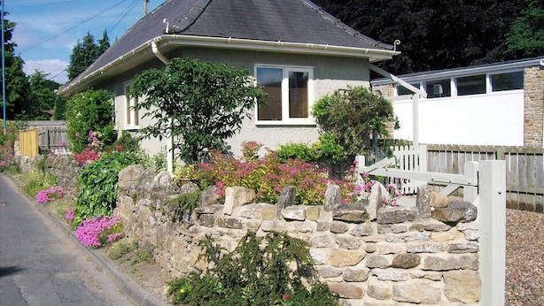 Une maison de plain-pied avec un mur en pierre, un petit jardin agrémenté de fleurs roses et une clôture blanche.