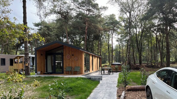 Una cabaña moderna con fachada de madera, un patio con mobiliario exterior y un coche aparcado cerca.