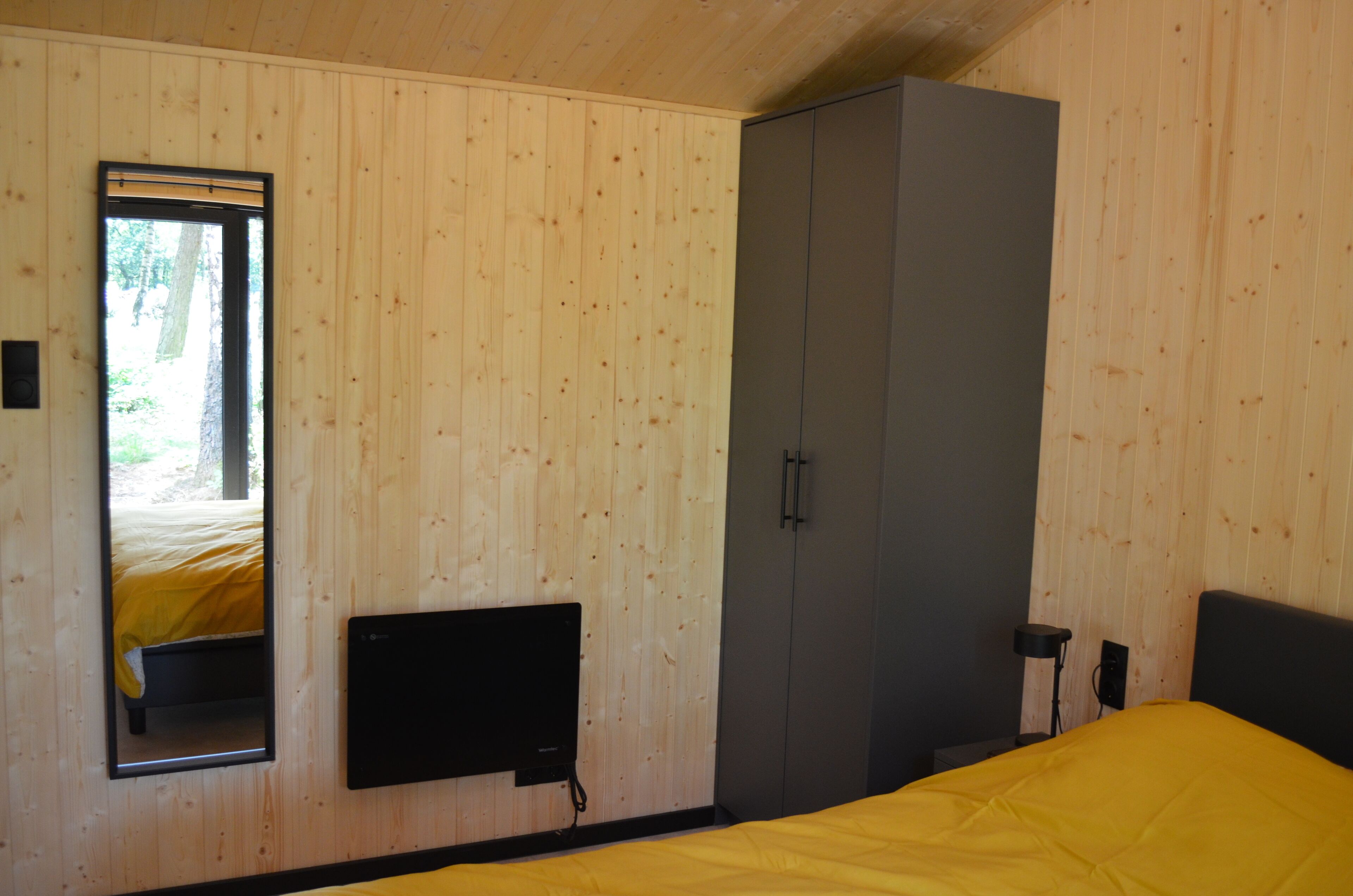 Une chambre avec des murs en bois, un lit recouvert d’une couverture jaune, une armoire, un téléviseur fixé au mur et une fenêtre donnant sur un paysage verdoyant.