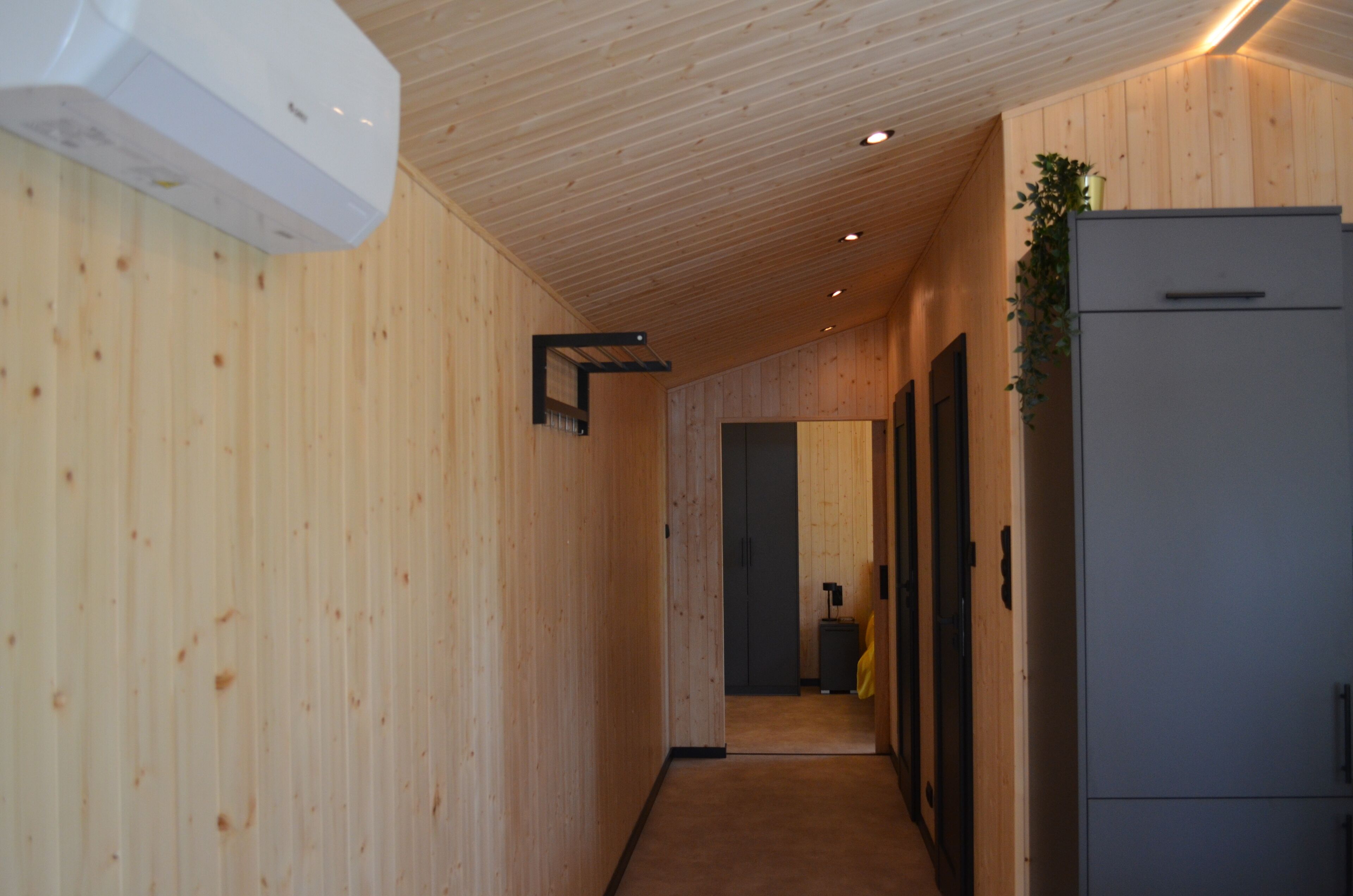 A narrow hallway with wooden walls and a ceiling, leading to a room with a refrigerator and a suitcase.
