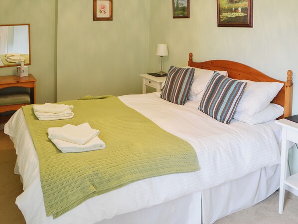 Cama bem arrumada com manta verde e almofadas brancas, mesa de cabeceira com abajur e quadro na parede.