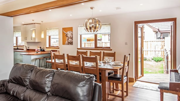 A modern living room with a leather sofa, wooden dining table, and chairs. There is a kitchen area with cabinets and a window.