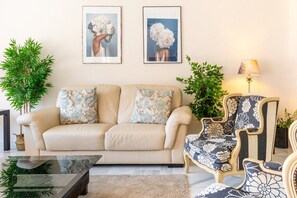 A living room with a beige sofa, floral cushions, a glass coffee table, a patterned armchair, and two framed pictures on the wall.