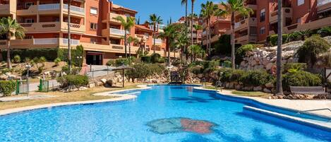 A swimming pool surrounded by multi-story buildings with balconies, palm trees, and a rocky landscape.