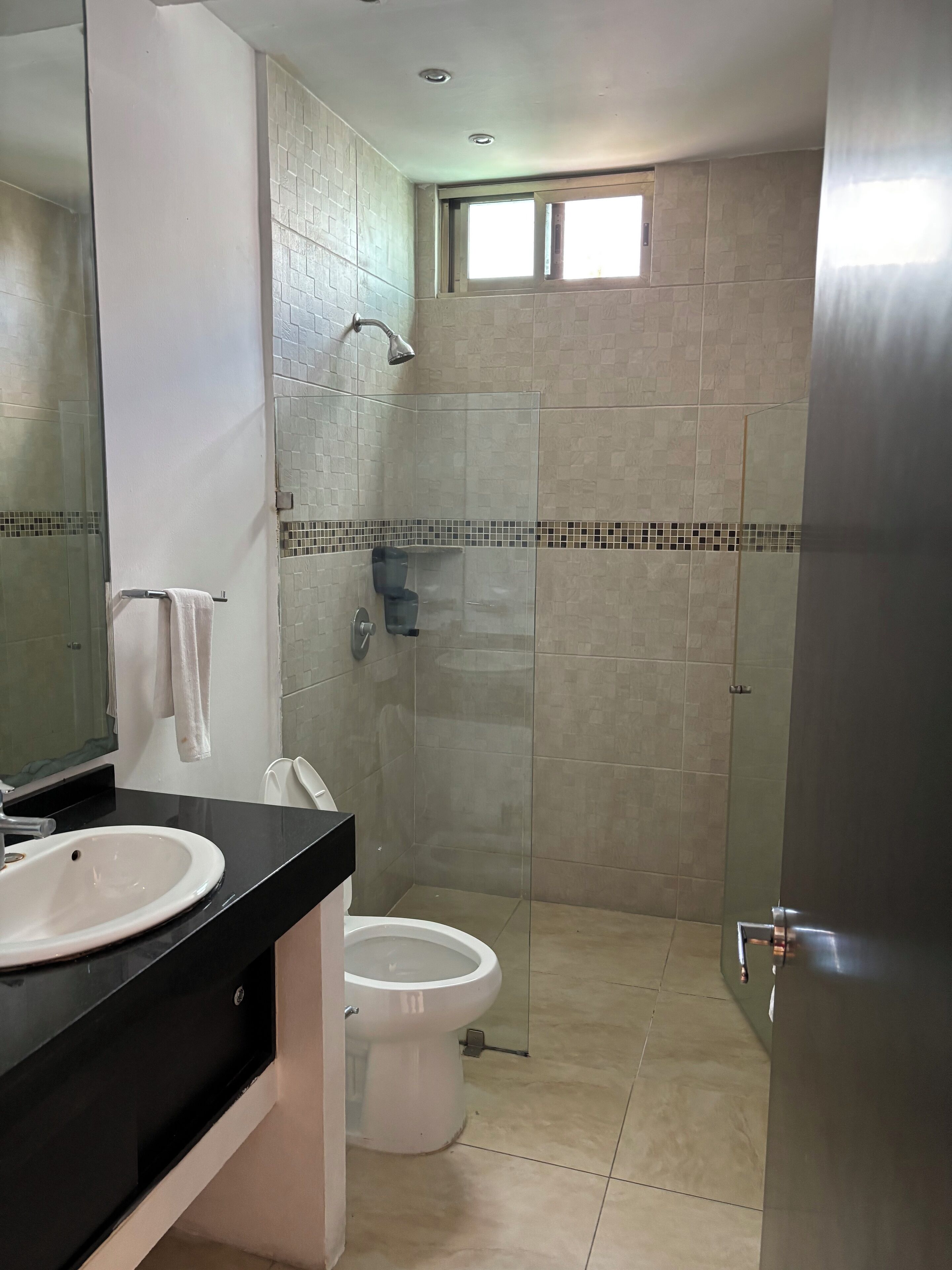 Un baño con un lavamanos blanco, un inodoro, una ducha con puerta de vidrio y un ventanal sobre la ducha.