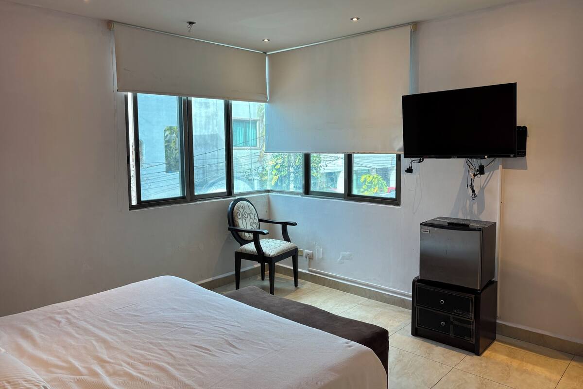 Una habitación de hotel moderna con una cama grande, una silla, un televisor montado en la pared y un armario pequeño.
