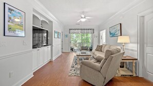 A spacious living room with a sofa, a coffee table, and a TV. There is a kitchen area with cabinets and a window. - Barefoot Havens 325 - Golf, Shopping, Dining, Pool (North Myrtle Beach)