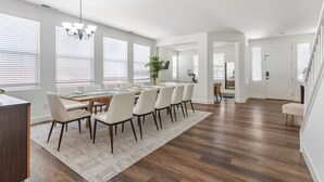 Une salle à manger spacieuse, avec une longue table et des chaises, un lustre et de grandes fenêtres équipées de stores.