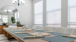 Une salle de conférence avec une longue table préparée pour une réunion, des chaises blanches, un lustre et de grandes fenêtres équipées de stores.