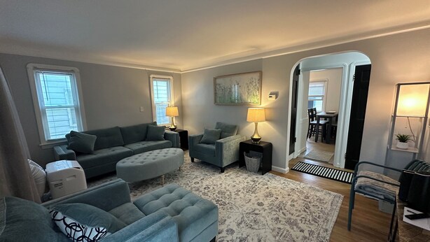 A living room with a grey sofa, ottoman, and armchair, a patterned rug, and a dining area visible through an archway.
