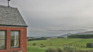 A stone building with a chimney, a window, and a view of green fields and distant hills.