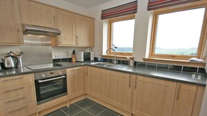 A modern kitchen with wooden cabinets, a stainless steel range hood, a microwave, and a built-in oven. There are two windows with striped curtains, a sink, and a tiled backsplash.