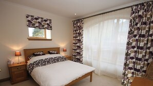 A bedroom with a bed, two bedside tables, a nightstand, a window with patterned curtains, and a view of the outdoors.