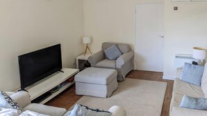 Una sala de estar con televisión de pantalla plana, un sofá, un reposapiés, una mesa de centro y una lámpara.