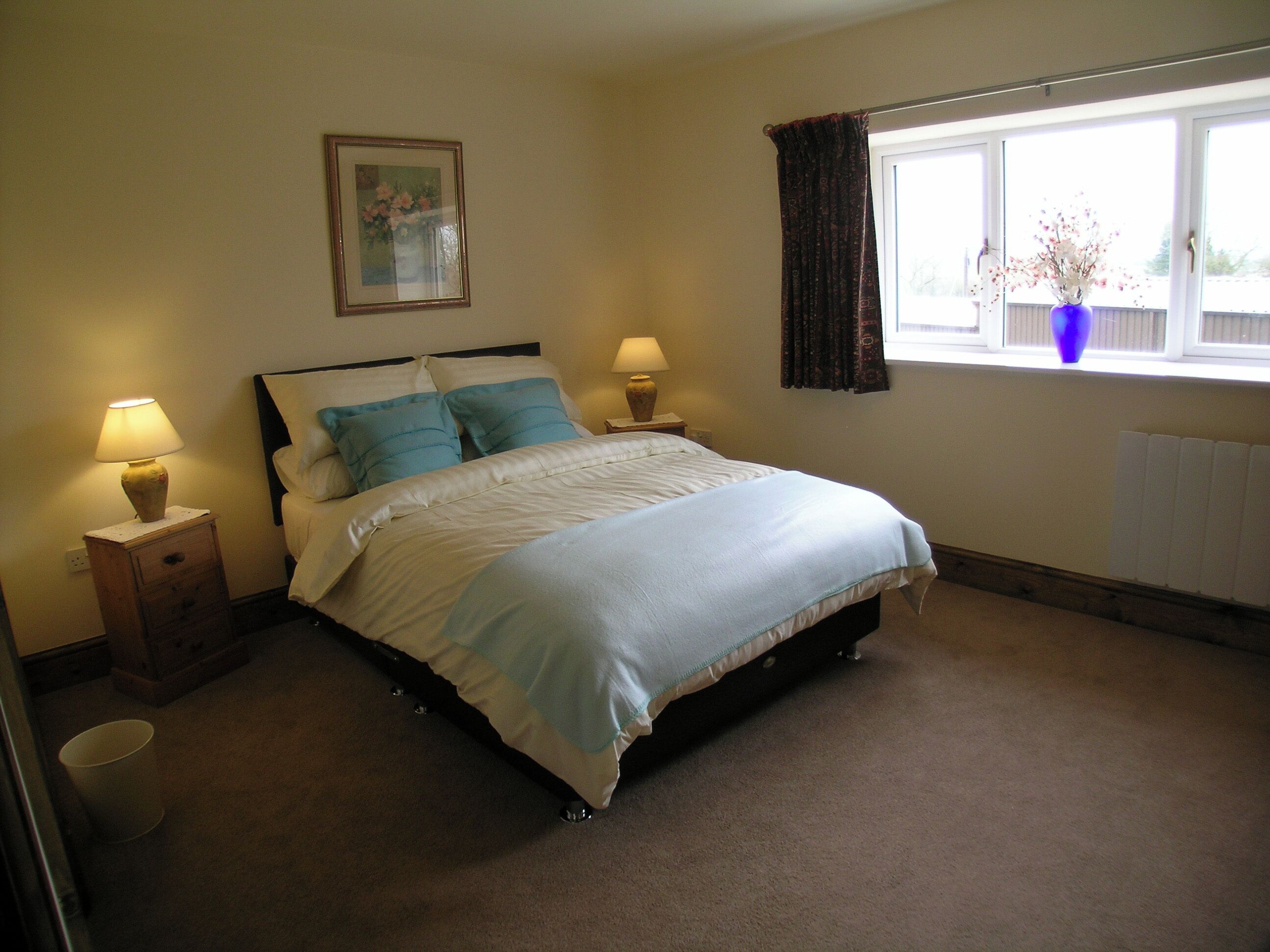 A bedroom with a bed, two bedside lamps, a nightstand, a window with curtains, and a wall-mounted picture.