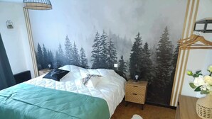Een slaapkamer met een bed, nachtkastje, lamp, een wanddecoratie van bomen, een houten dressoir en een witte vloerkleed.