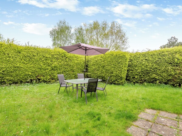 A patio with a table and chairs, an umbrella, and a hedge in the background.