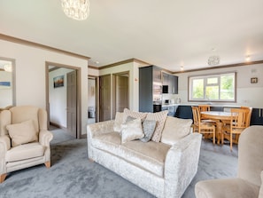 A living room with a beige sofa, a wooden dining table with chairs, a kitchen area with cabinets and a window, and a hallway with a door.