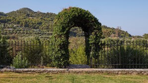 Un arco cubierto de vegetación, un sendero de piedra y una cerca metálica en un área ajardinada, con una montaña al fondo.