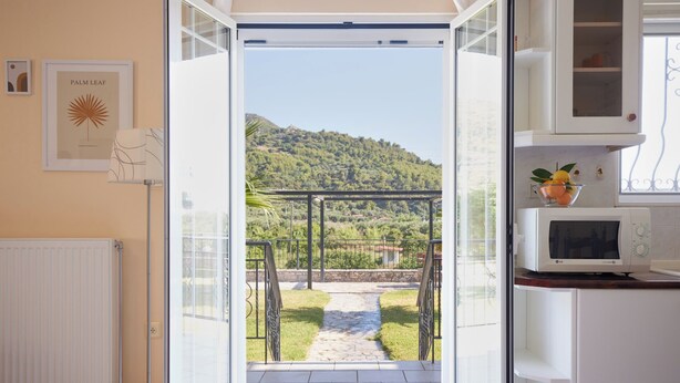 Puerta abierta que da a un balcón con vistas a una colina, un radiador blanco y una zona de cocina con microondas y frutero.