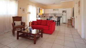 Una sala de estar con un sofá rojo, una mesa de centro de madera y un sillón de mimbre. Se observa una cocina al fondo con armarios blancos y un refrigerador.