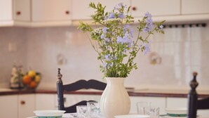Una mesa de comedor con platos y vasos blancos, un jarrón blanco con flores azules, y una silla con respaldo de madera oscura.