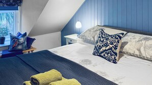 Une chambre avec un plafond en pente, un lit agrémenté d’oreillers à motifs, une table de chevet avec une lampe et une fenêtre donnant sur des arbres.