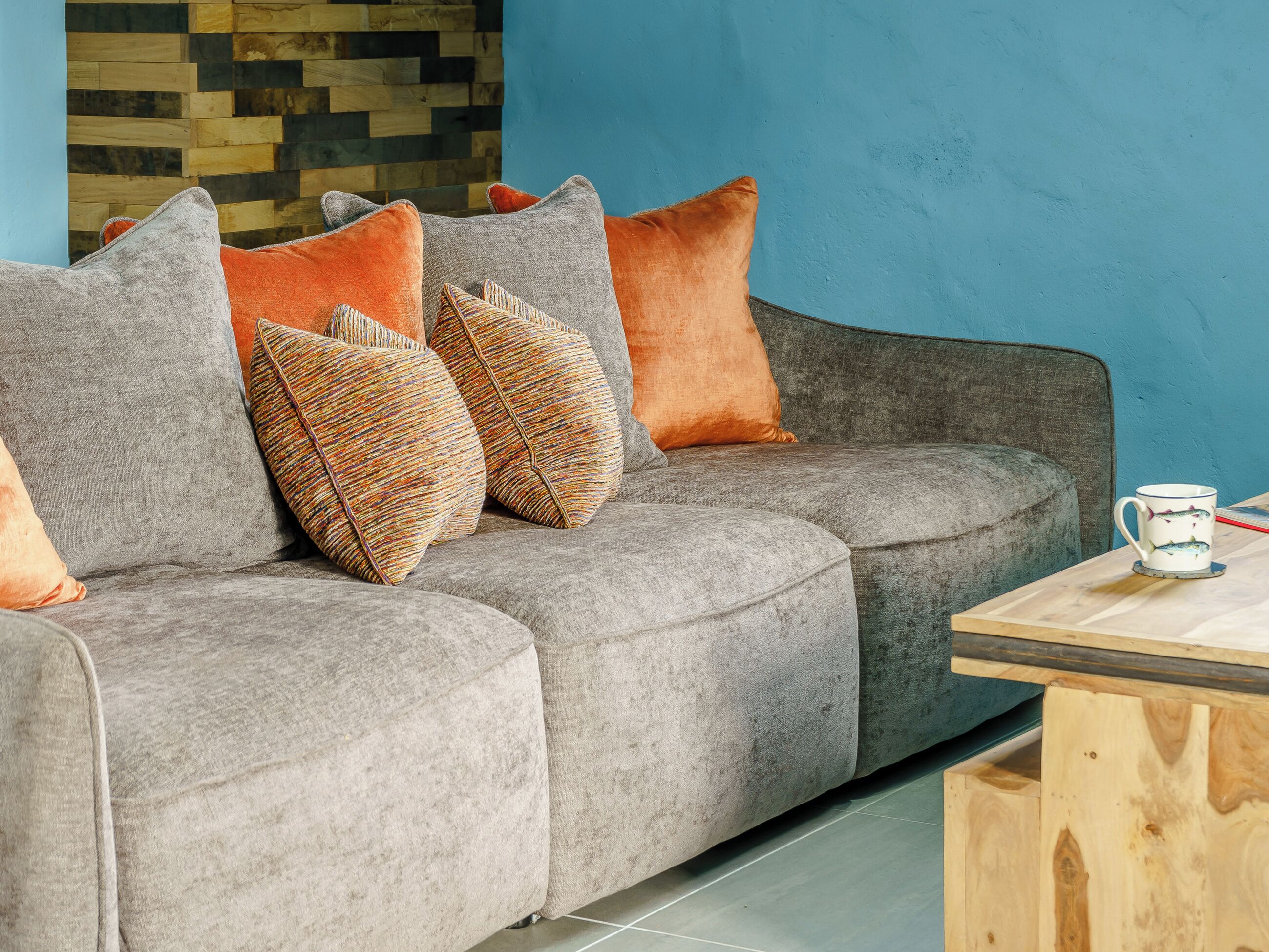 Un canapé gris agrémenté de plusieurs coussins, une table basse en bois et un mur bleu orné d’un motif en lambris.