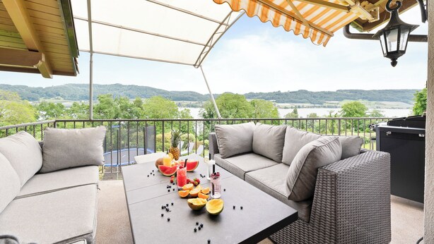 Ein Balkon mit Sitzgelegenheit im Freien, einem Tisch mit Obst und Getränken sowie Blick auf einen See und Hügel.