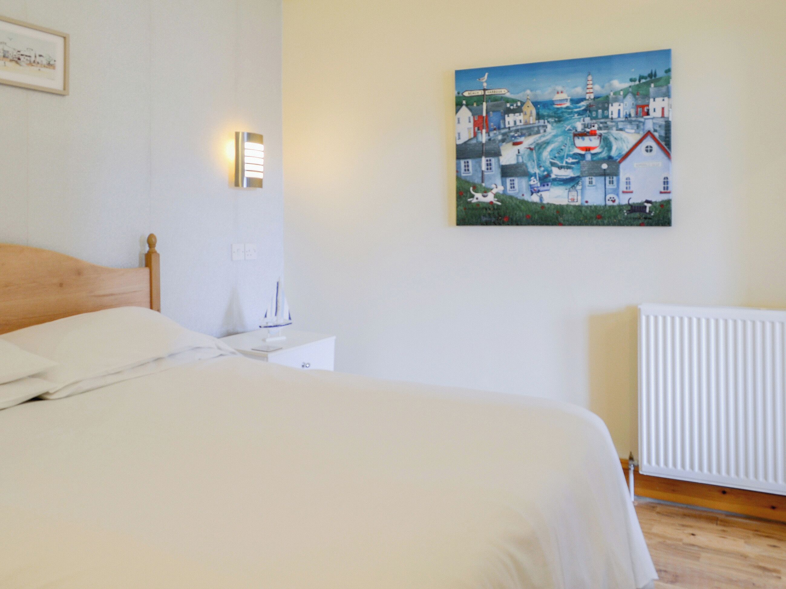 A neatly made bed in a room with a painting of a coastal town on the wall, a bedside table with a lamp, and a radiator.