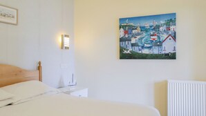 Una cama bien hecha en una habitación con un cuadro de una ciudad costera en la pared, una mesita de noche con una lámpara y un radiador.