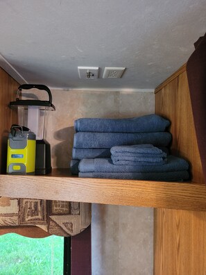 A compact room with a wooden shelf holding a water purifier, a kettle, and folded towels.