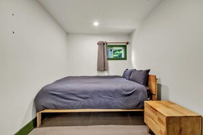 A bedroom with a single bed, a wooden nightstand, a small window with a view of greenery, and a curtain. - White Birches Cottages (Dwight)