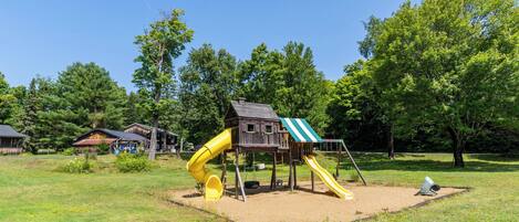 A playground with a slide and swings, surrounded by trees and a grassy area.