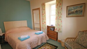 Ein Schlafzimmer mit Bett, Nachttisch, Bücherregal, Sessel und Fenster mit Vorhängen.