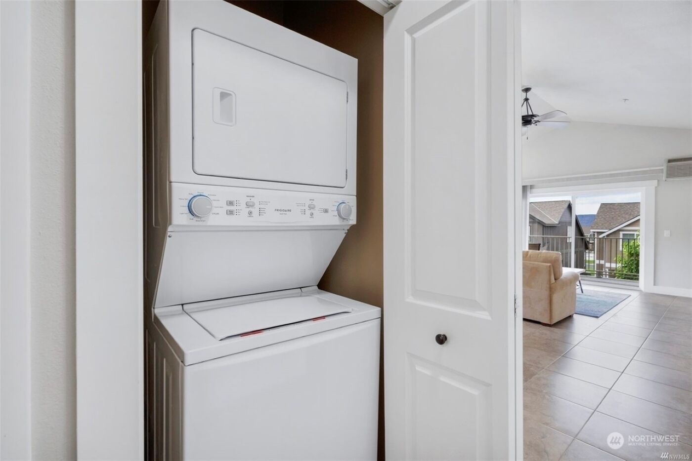 Une buanderie équipée d’une machine à laver et d’un sèche-linge superposés, une porte donnant sur un espace de vie avec un canapé et une vue sur un quartier résidentiel.