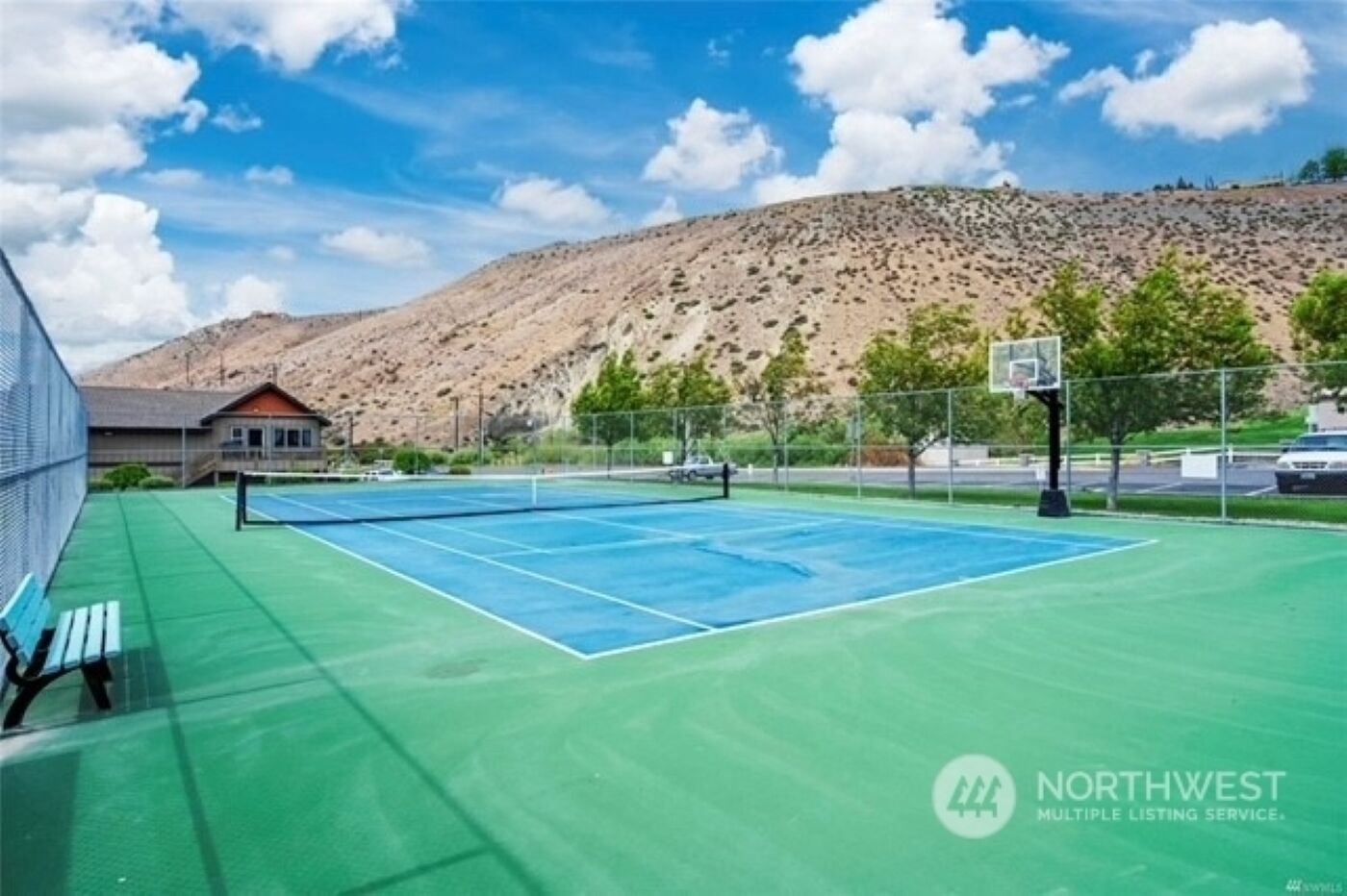 An outdoor tennis court with a green surface, a basketball hoop, and a bench.