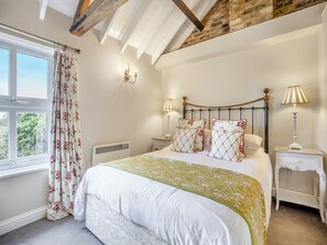A bedroom with a bed, two bedside tables, a radiator, a window with curtains, and a sloped ceiling.