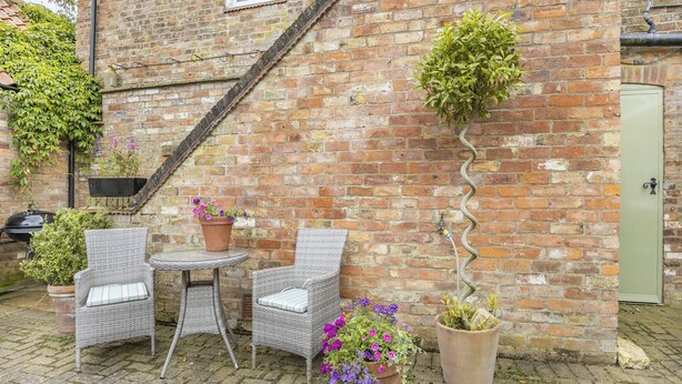 A brick building with a small patio area featuring wicker chairs, a round table, and potted plants.