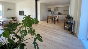 An open-plan living and dining area with a wooden floor, a dining table with chairs, a kitchen area with appliances, and a large potted plant in the foreground.