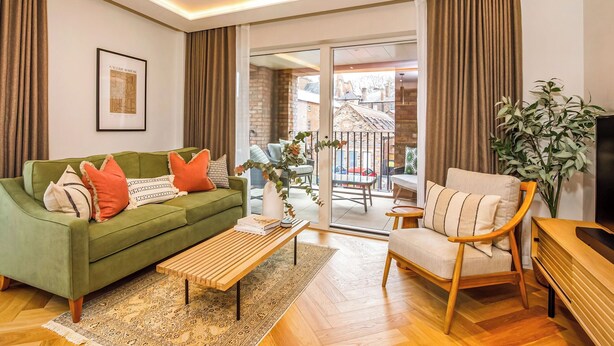 A modern living room with a green sofa, a wooden coffee table, and a beige armchair. There is a framed picture on the wall, a potted plant, and a view of the outside through a glass door.