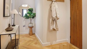 A hallway with a wooden floor, a coat rack with a coat, a potted plant, a mirror, and a framed picture on the wall.