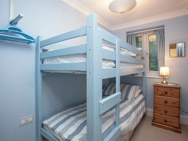 A bunk bed with a striped mattress, a wooden dresser with drawers, a lamp, and a window with curtains.