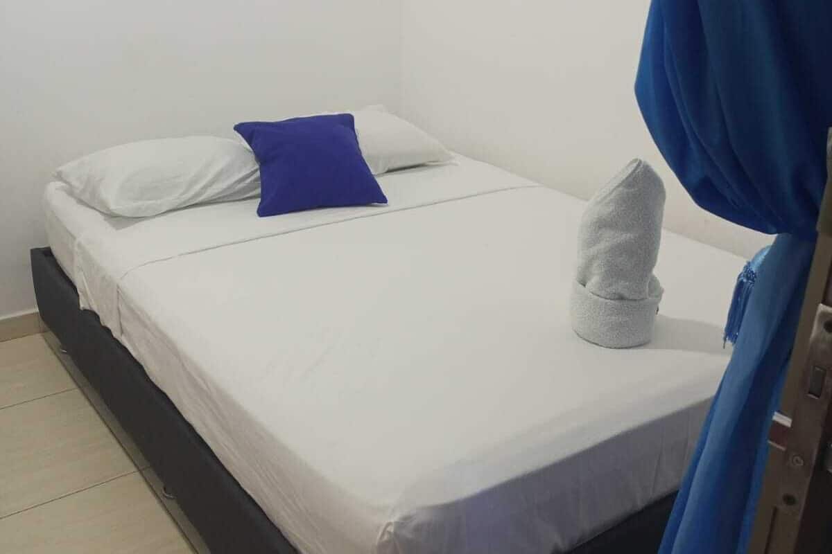 Una cama individual con ropa de cama blanca y una almohada azul, una toalla sobre la cama y una cortina azul parcialmente visible.