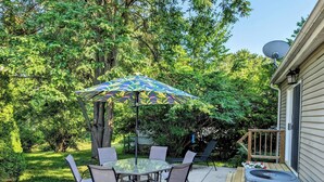 Een terras buiten met een glazen tafel en stoelen, een paraplu met een patroon en een schotelantenne op het huis.